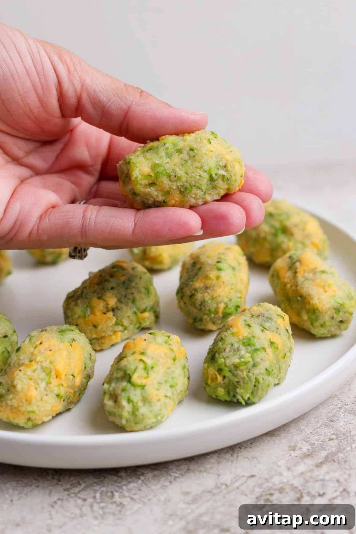 Crispy Air Fryer Zucchini Tots 5 A hand holds a homemade broccoli and cheese tot over a white plate with more tots arranged on it. The tot is golden and speckled with green.