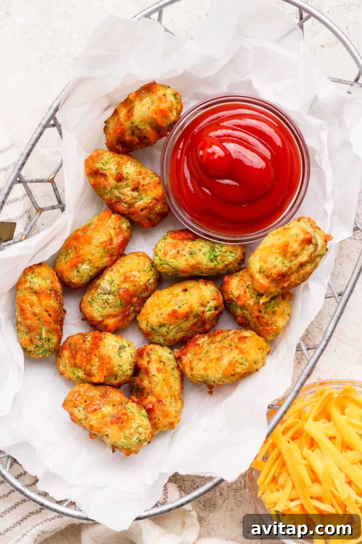 Crispy Air Fryer Zucchini Tots 6 A basket of baked vegetable tots on parchment paper served with a bowl of ketchup; shredded cheese is visible beside the basket.