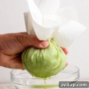 A hand squeezes shredded green zucchini wrapped in paper towels over a glass bowl, extracting liquid.