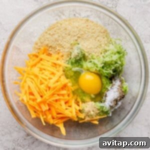 A glass bowl containing shredded cheddar cheese, grated zucchini, an egg, breadcrumbs, and seasonings on a light textured surface, ready to be mixed.