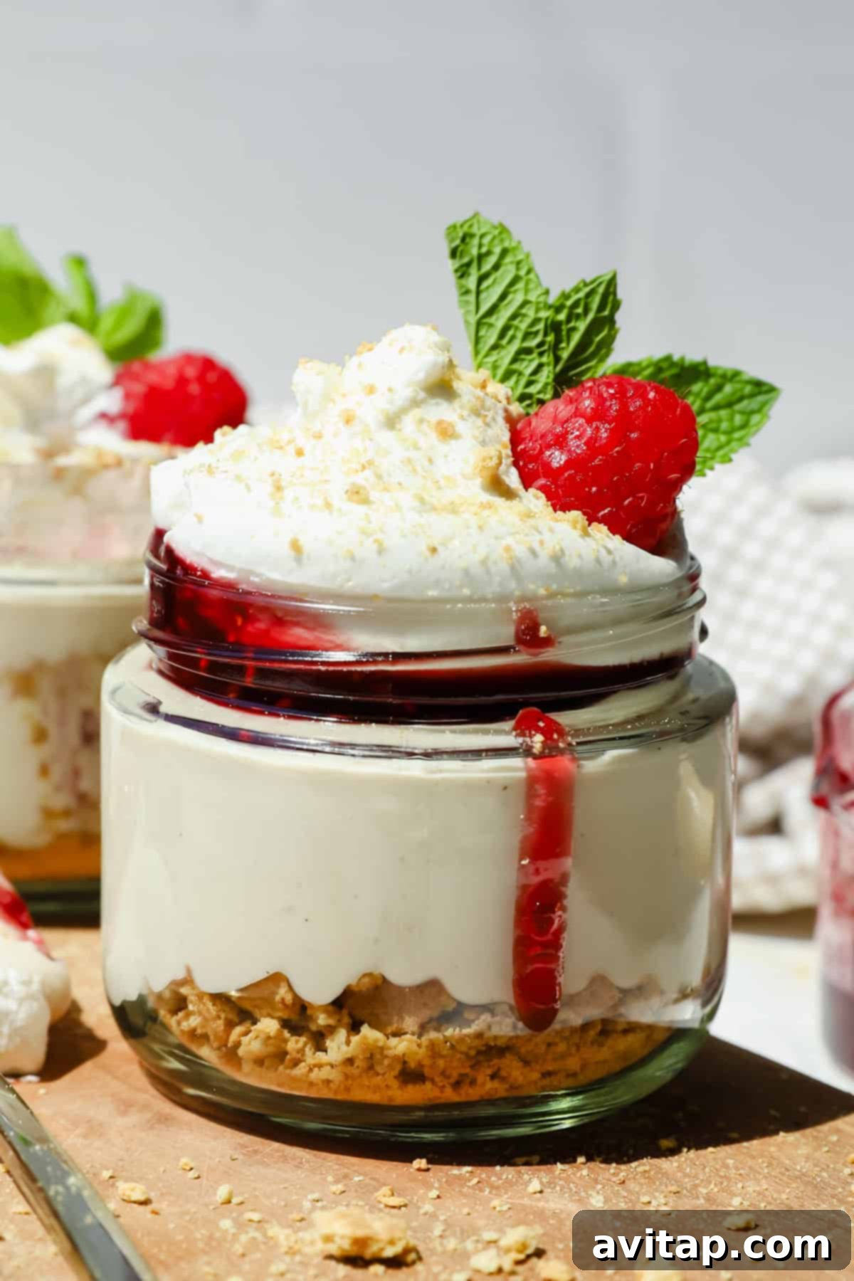 Guilt-Free Protein Cheesecake Jars 2 A small glass jar of cheesecake topped with whipped cream, raspberry sauce, a fresh raspberry, and mint leaves, with a layer of crushed biscuits at the bottom.