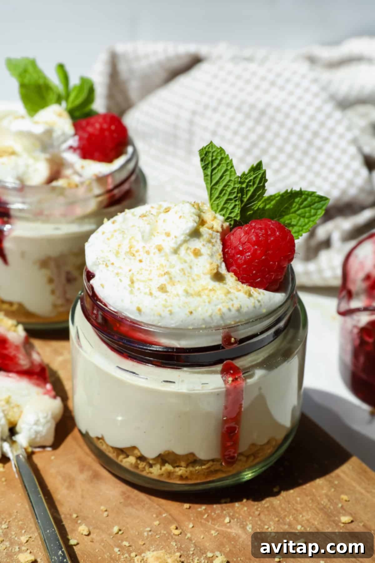 Guilt-Free Protein Cheesecake Jars 4 A glass jar filled with layered cheesecake, graham cracker crust, whipped cream, a raspberry, and mint leaves, with berry sauce dripping down the side.