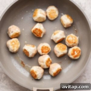 A beige skillet contains partially cooked, lightly browned meatballs spread out on its surface.