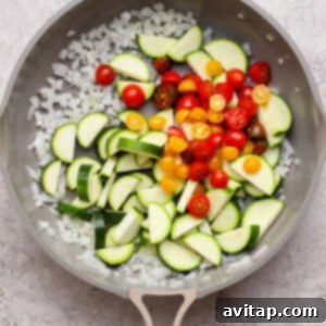 A frying pan contains chopped onions, sliced zucchini, and halved cherry tomatoes on a light textured surface.