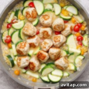A skillet with browned meatballs, sliced zucchini, yellow squash, cherry tomatoes, and orzo in a light sauce on a gray surface.