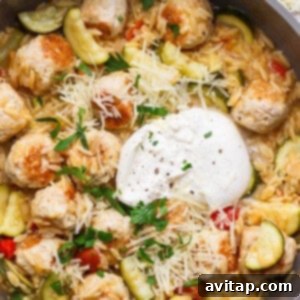 A skillet filled with orzo pasta, zucchini, meatballs, diced tomatoes, shredded cheese, a dollop of sour cream, and chopped parsley.