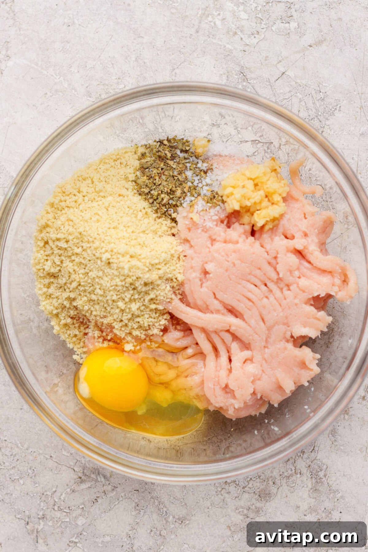 Ingredients for chicken meatballs including ground chicken, a cracked egg, breadcrumbs, minced garlic, salt, and dried herbs in a glass bowl.