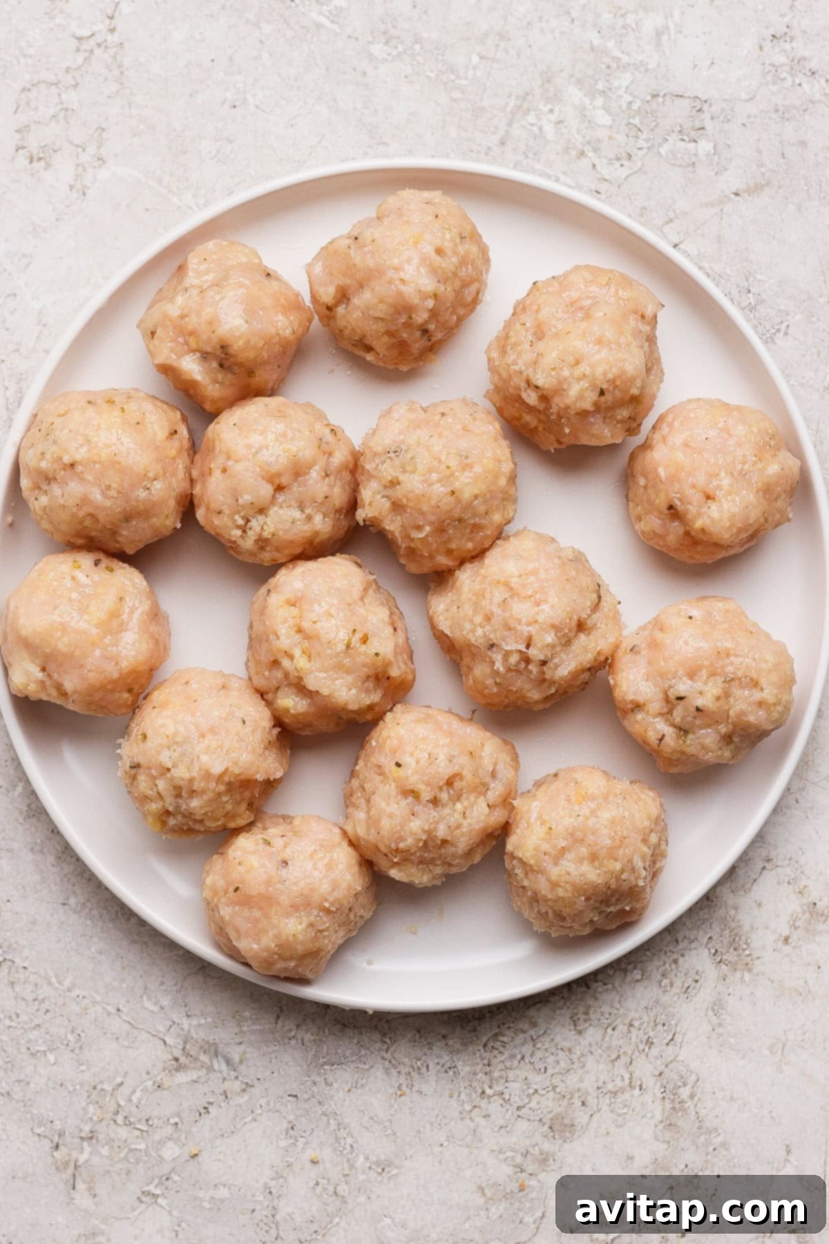 Sixteen perfectly formed raw chicken meatballs arranged neatly in a circular pattern on a white plate, ready for cooking.