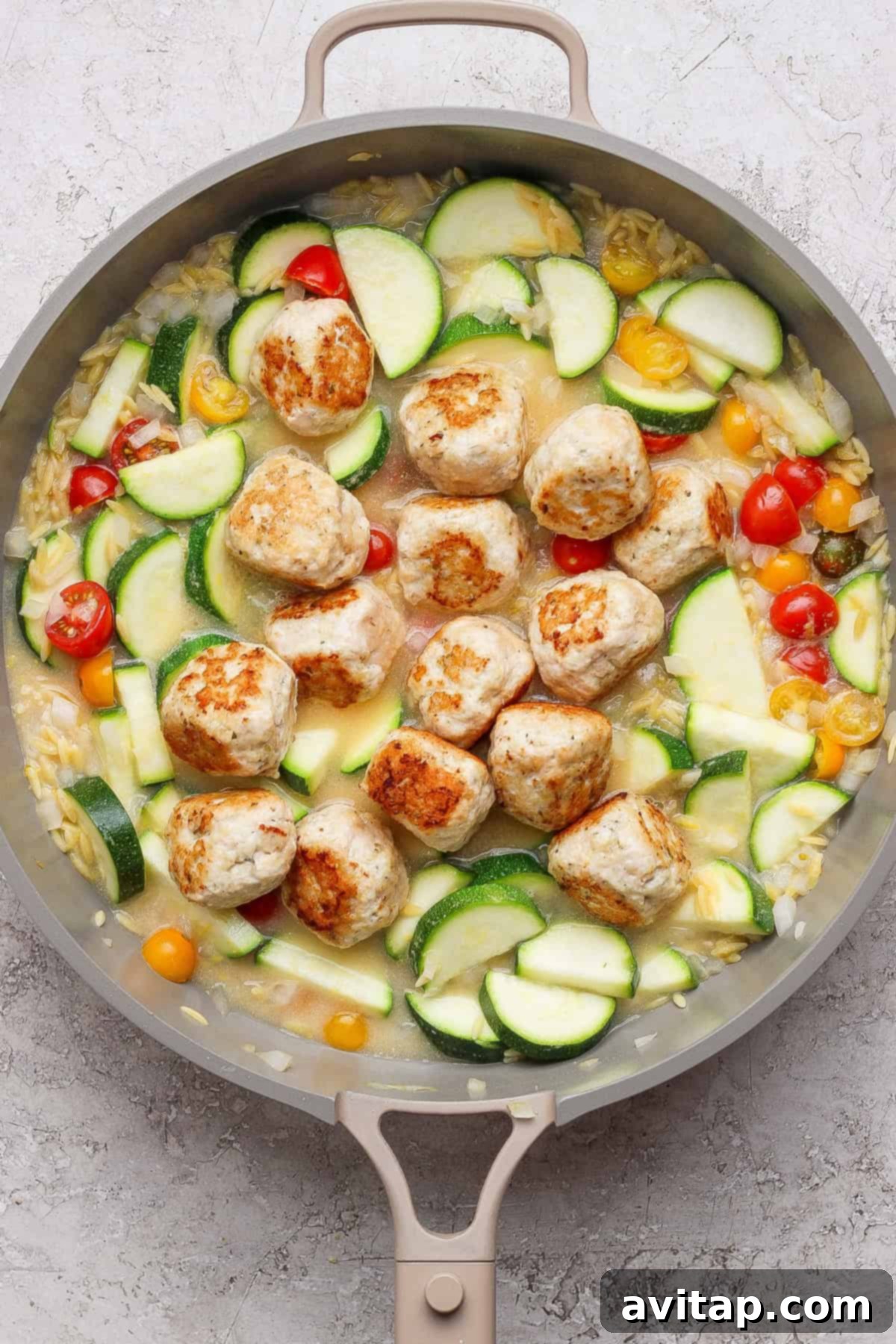 A skillet showcasing browned chicken meatballs, sliced zucchini, yellow squash, cherry tomatoes, and orzo pasta simmering in a light, flavorful sauce.