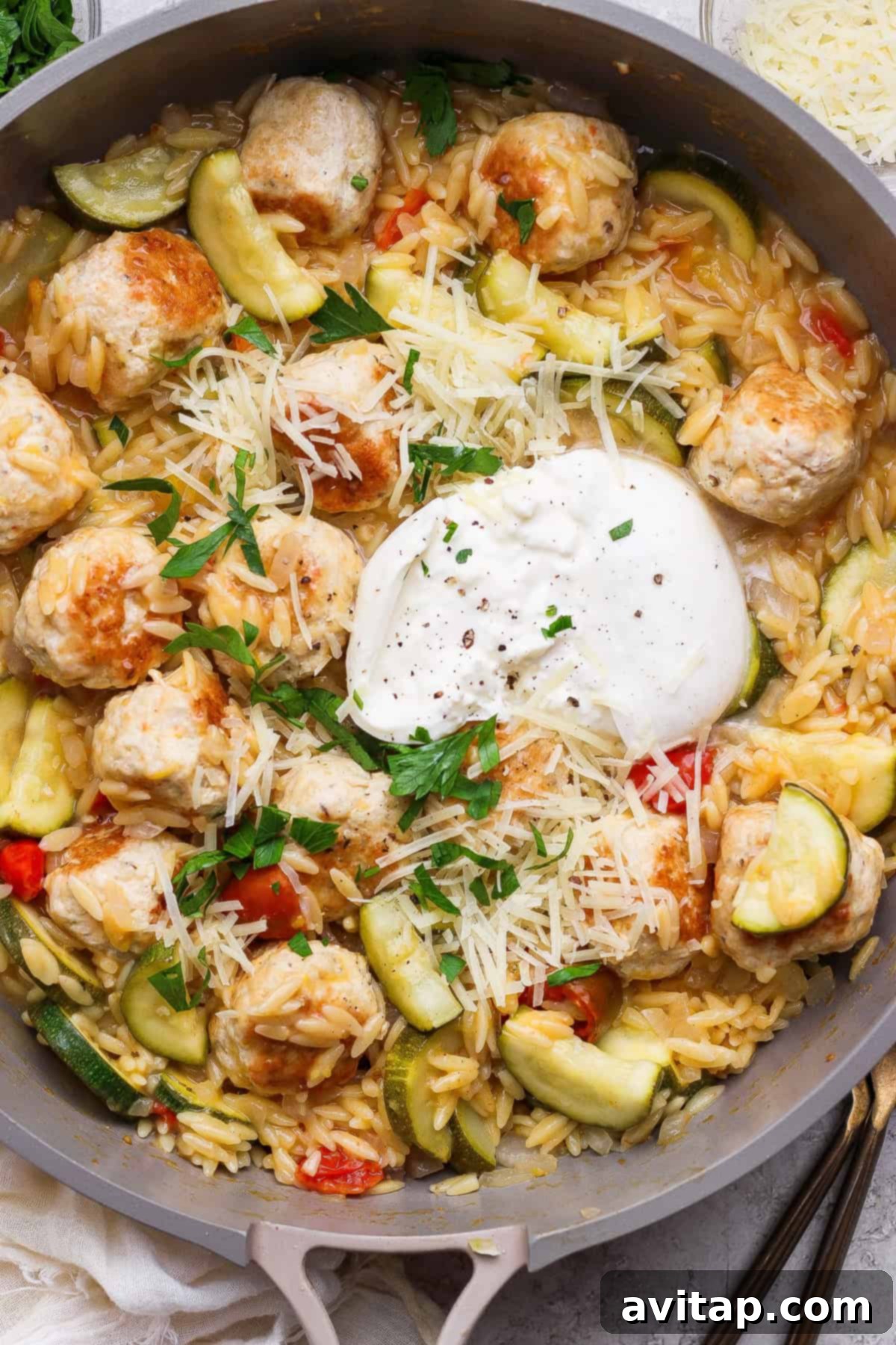 A beautifully presented skillet dish featuring orzo pasta, golden-brown chicken meatballs, vibrant zucchini slices, juicy cherry tomatoes, garnished with fresh parsley, grated Parmesan cheese, and a luxurious melted burrata in the center, ready to be enjoyed.