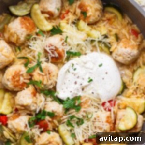 A skillet filled with orzo pasta, zucchini, meatballs, diced tomatoes, shredded cheese, a dollop of sour cream, and chopped parsley.
