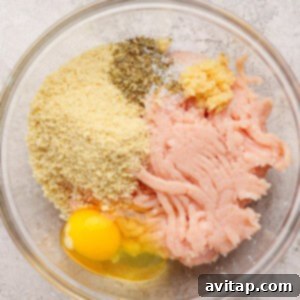 A glass bowl containing ground chicken, a cracked egg, breadcrumbs, minced garlic, salt, and dried herbs on a light textured surface.