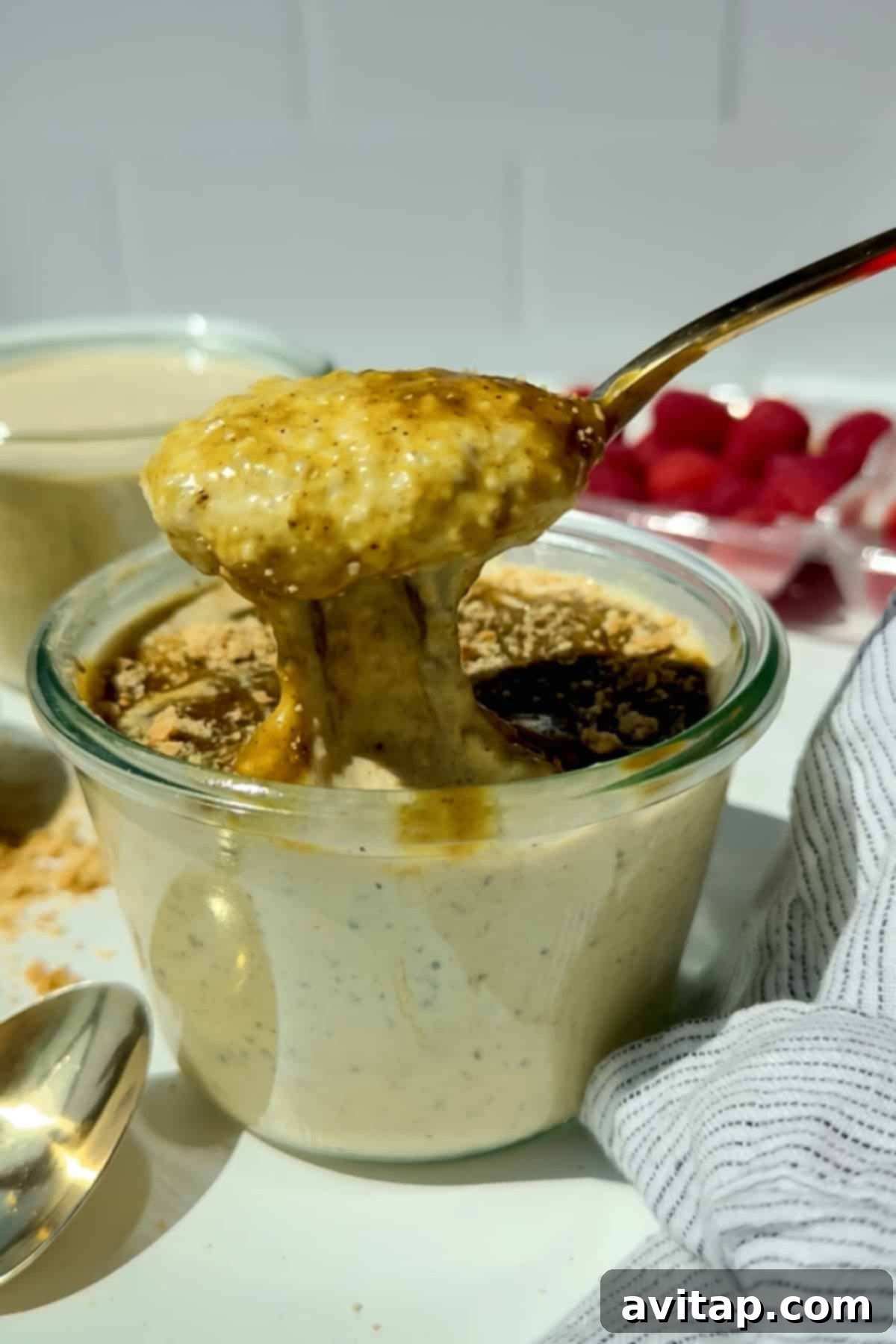 A spoon lifts creamy, blended pistachio overnight oats from a glass jar, topped with a drizzle of syrup and a sprinkle of pistachio crumbs, with fresh raspberries and a striped cloth nearby.