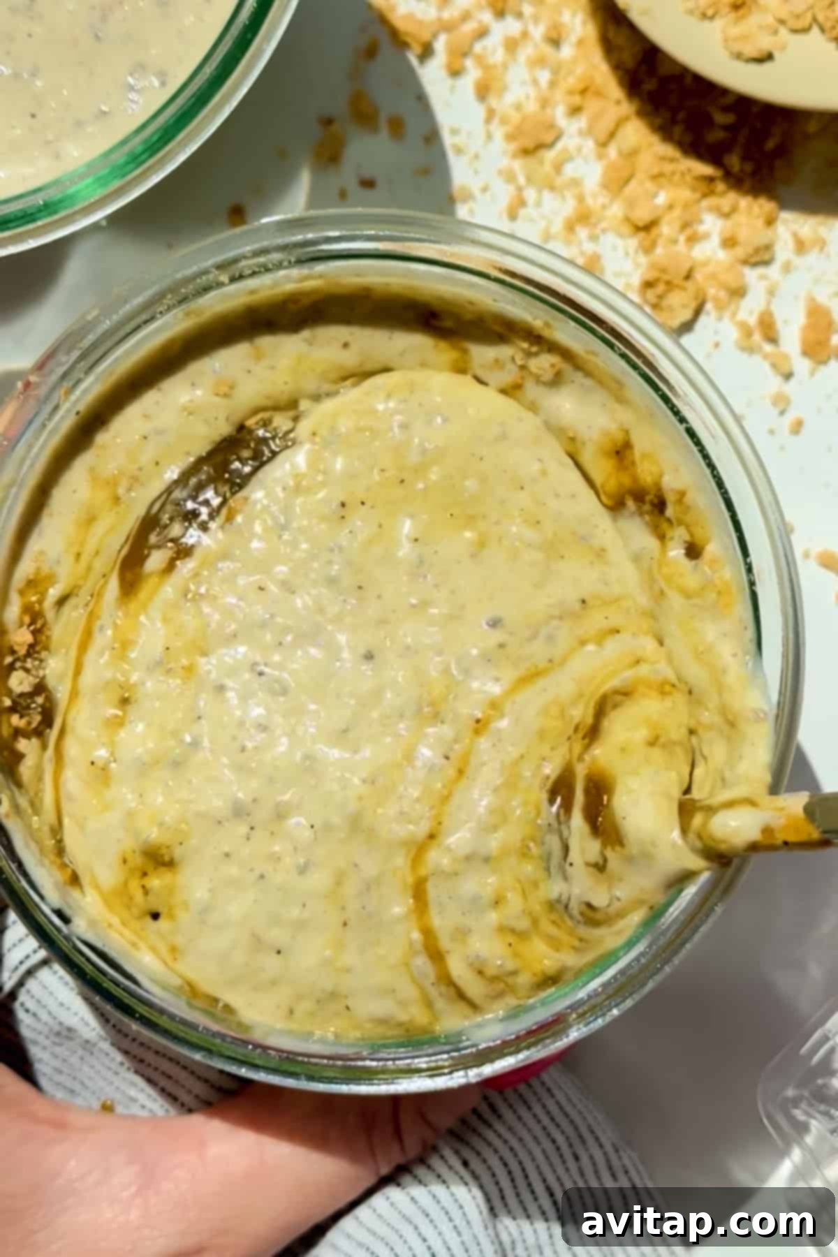 A glass bowl containing a creamy, beige pistachio oat mixture, being stirred with a spoon, highlighting its smooth and luxurious texture. Crumbled pistachio pieces are scattered nearby on a clean white surface.