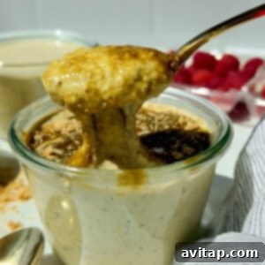 A spoon lifts thick, creamy pistachio pudding from a glass jar, with crumbs and a dark sauce on top; fresh raspberries are visible in the background, enticing viewers to try this delightful breakfast.