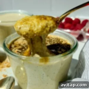 A spoon lifts thick, creamy pudding from a glass jar, with crumbs and a dark sauce on top; fresh raspberries are visible in the background.