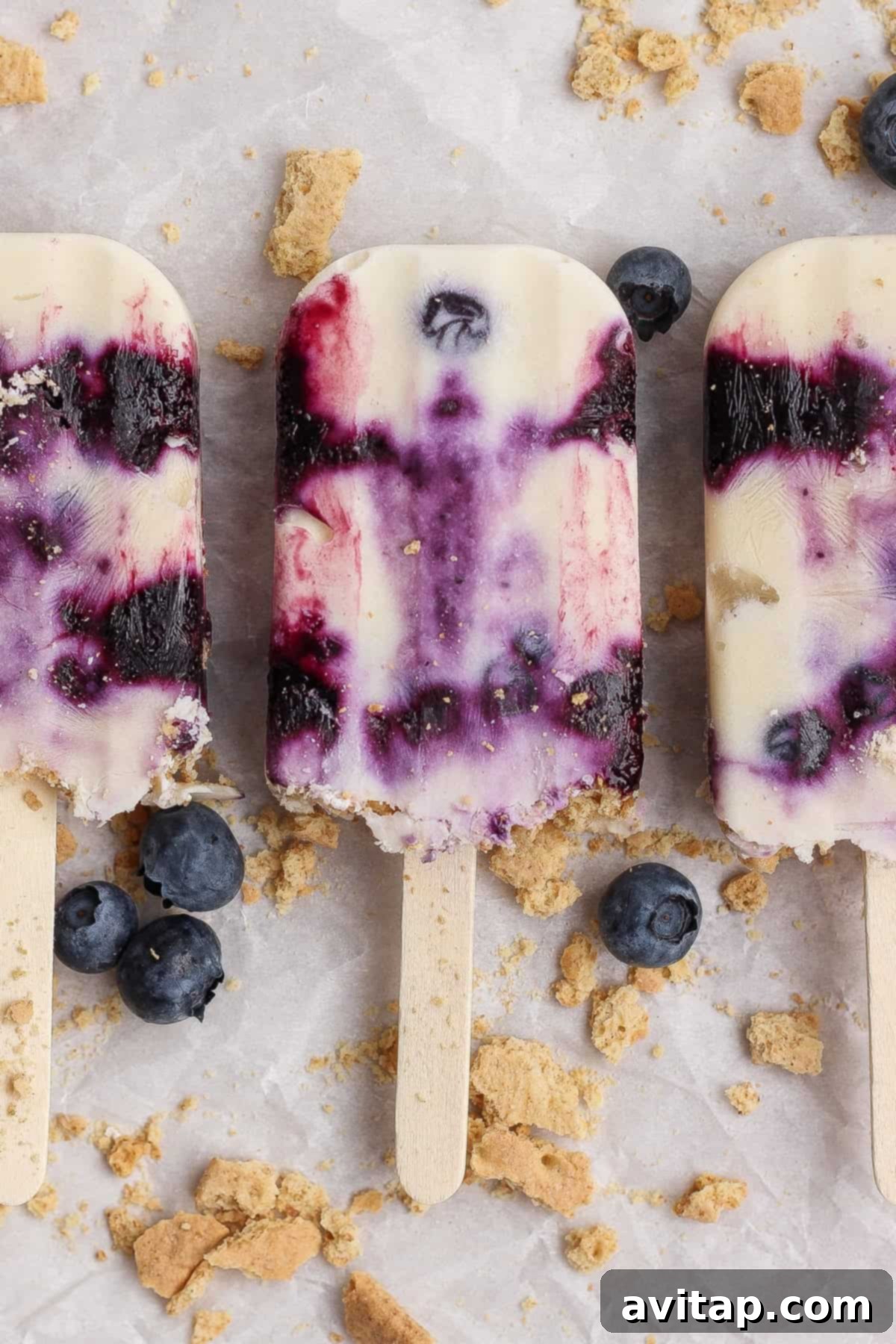 Three beautifully arranged blueberry cheesecake popsicles, showcasing visible blueberry swirls and creamy white layers, on a surface adorned with fresh blueberries and crumbled graham crackers.