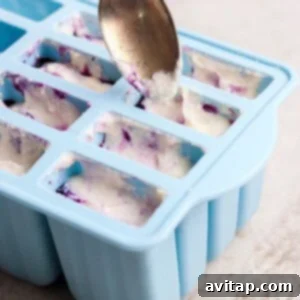 A spoon meticulously fills a blue silicone mold tray, layering a white creamy mixture with a purple fruit sauce to create a beautiful swirled effect.