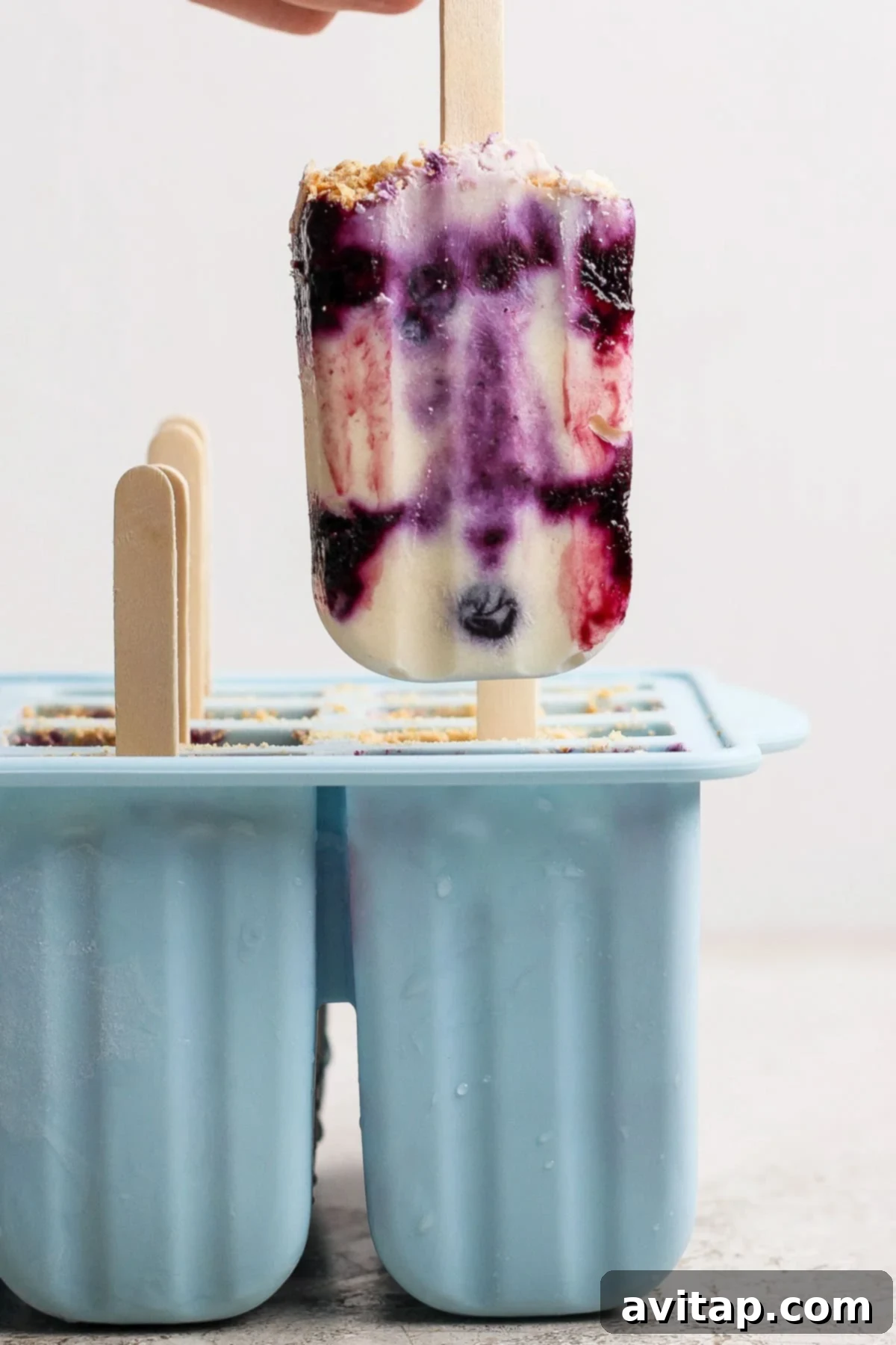 A hand holds a vibrant blueberry cheesecake popsicle with distinct purple, white, and red swirls, positioned above a blue silicone popsicle mold containing several more frozen popsicles.