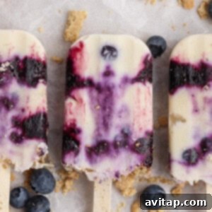 Three creamy blueberry popsicles with beautiful purple swirls are artistically arranged on a light-colored surface, surrounded by scattered fresh blueberries and delicate crumbled cookie pieces.