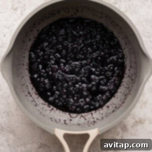 A beige saucepan filled with cooked blueberries simmering in a thick purple sauce, indicating the jam preparation stage.