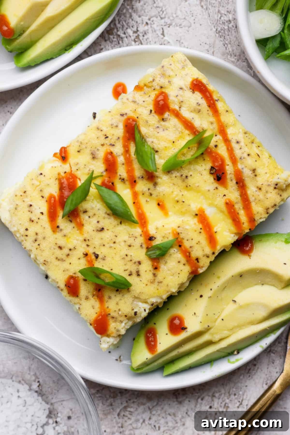 A vibrant rectangular sheet pan egg dish, generously topped with fresh green onions and a drizzle of hot sauce, served alongside creamy avocado slices on a pristine white plate, highlighting a perfect breakfast meal.