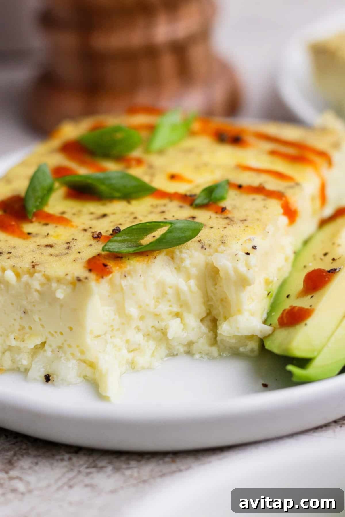 A generous slice of fluffy baked egg casserole is presented on a white plate, elegantly garnished with a sprinkle of fresh green onions and a bold swirl of hot sauce, accompanied by neatly fanned avocado slices.