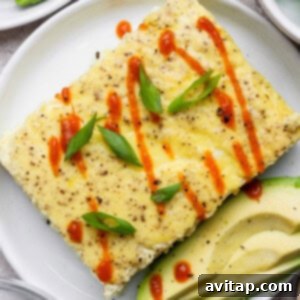 A rectangular egg dish garnished with sliced green onions and hot sauce, served with avocado slices on a white plate.