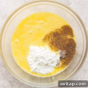 A glass bowl containing whisked eggs, cottage cheese, ground black pepper, and salt on a light textured surface, ready for baking.