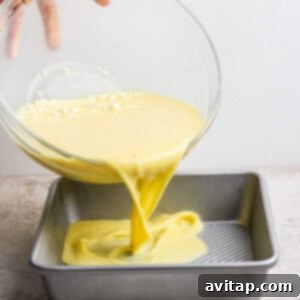 A hand pours the yellow egg batter from a clear glass bowl into a square metal baking pan, situated on a kitchen countertop.