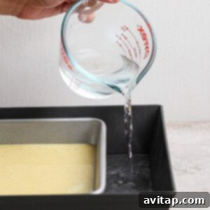 A hand carefully pours water from a glass measuring cup into a larger baking pan, which cradles a smaller pan filled with an egg mixture, setting up the water bath for baking.