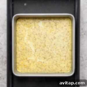 A square metal baking pan filled with a yellow, speckled egg mixture rests inside a larger black tray, positioned on a textured gray surface, showcasing the eggs baking in a water bath.