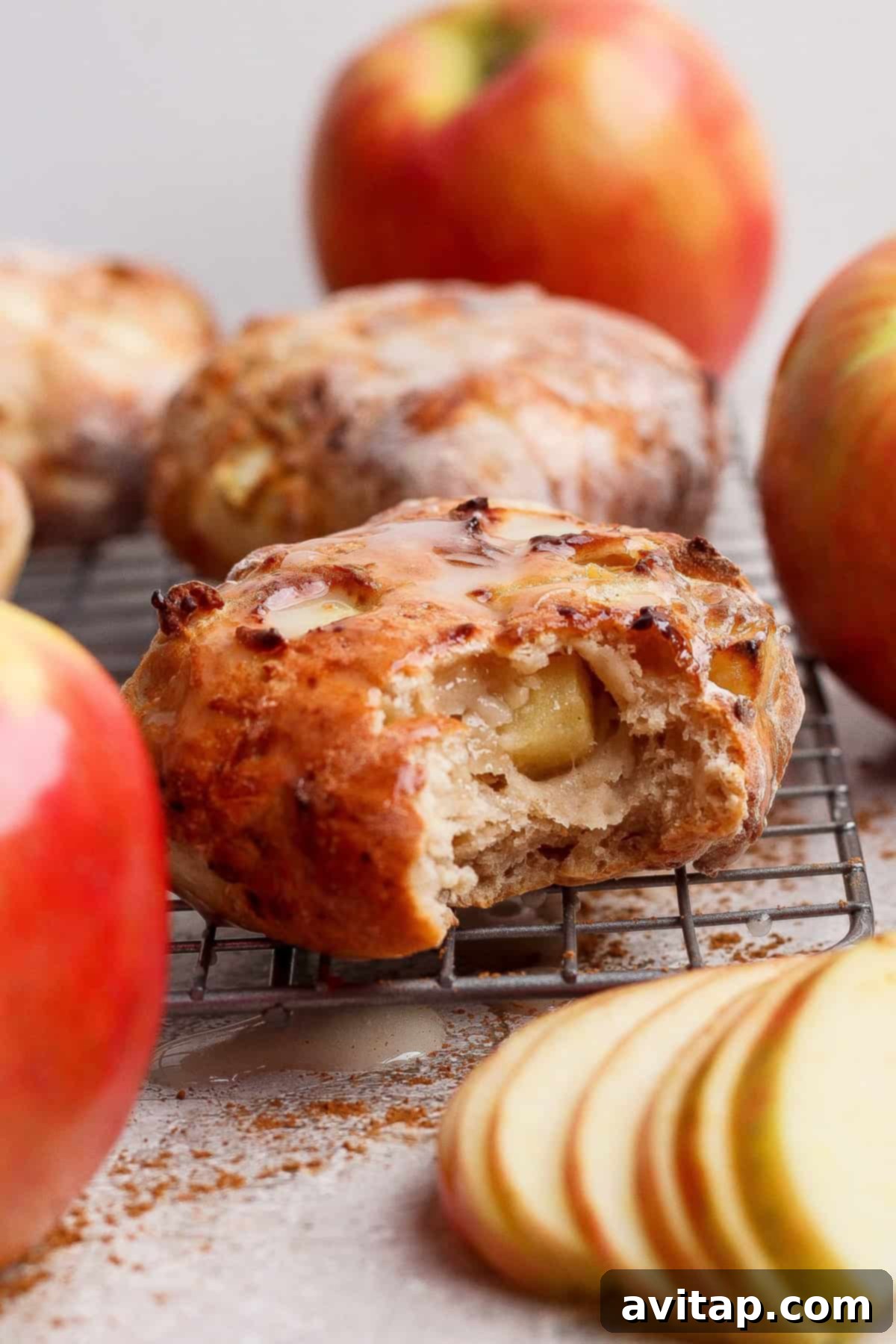 A partially eaten air-fried apple fritter sits on a cooling rack, surrounded by whole apples and apple slices, showcasing its delightful texture and the freshness of the ingredients.