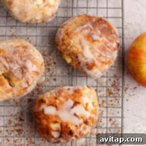 Four apple-studded pastries with glaze sit on a cooling rack, surrounded by apples, cinnamon, and apple slices on a light surface.