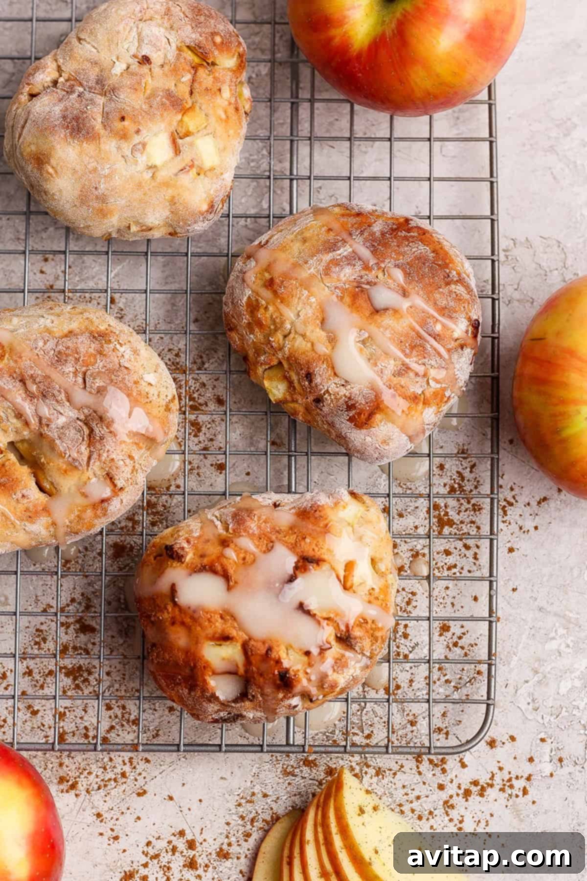 Four perfectly glazed apple-studded pastries sit on a cooling rack, surrounded by fresh apples, ground cinnamon, and apple slices on a light surface, ready to be enjoyed.