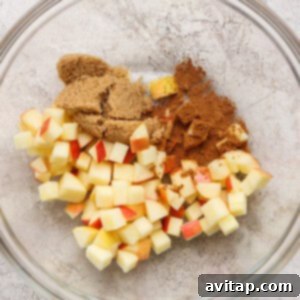 A glass bowl containing diced apples, brown sugar, and ground cinnamon on a light textured surface.