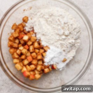 A glass bowl with diced apples coated in cinnamon on one side and a mixture of flour and white powder, likely baking powder, on the other side, on a light textured surface.