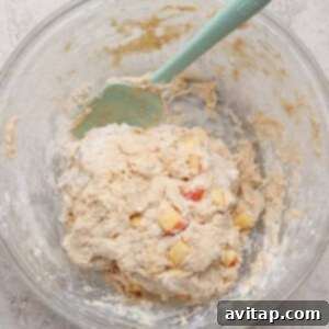A glass bowl containing dough mixed with small apple chunks and a green spatula on a light textured surface.