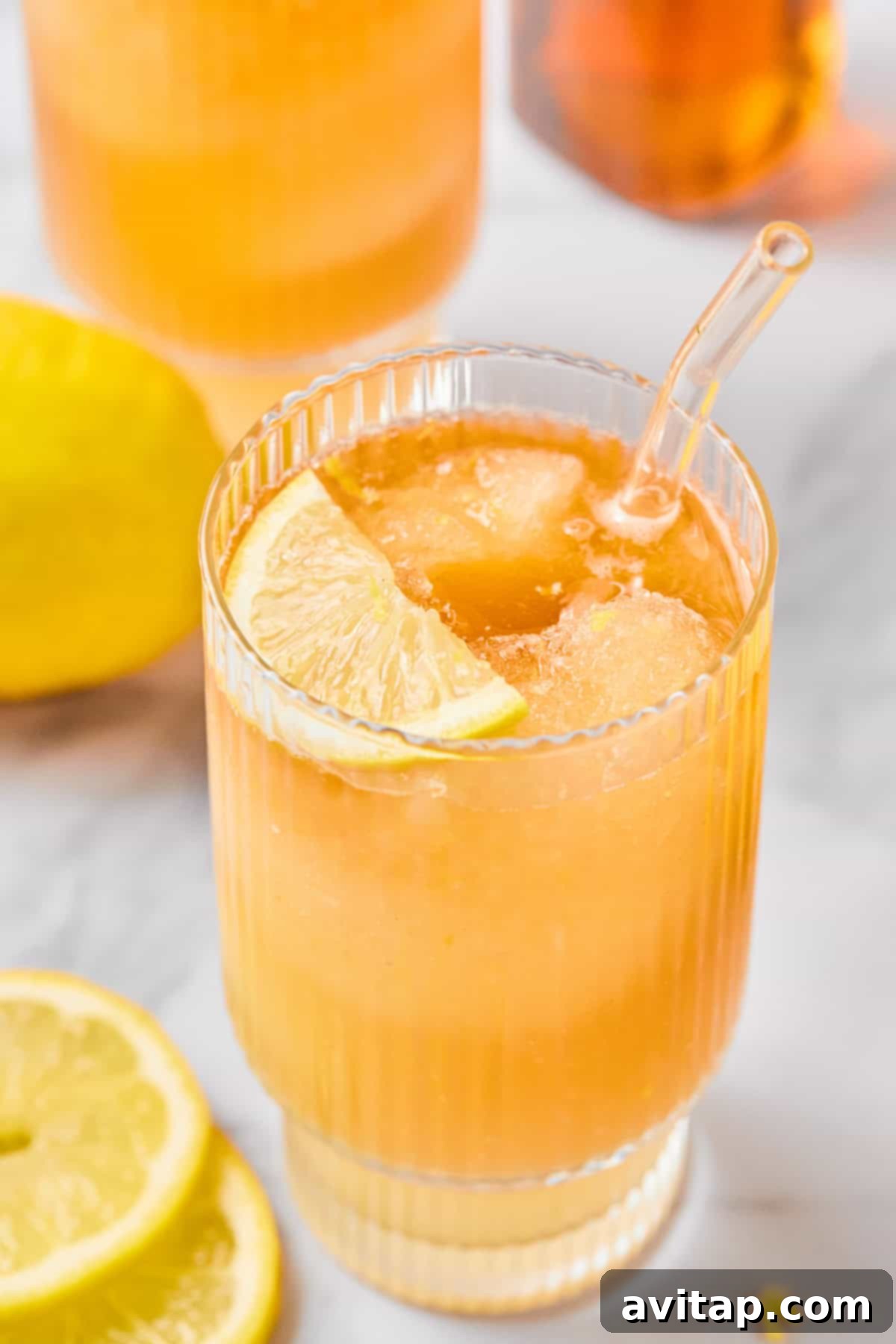 A vibrant glass of iced lemon drink, garnished with a fresh lemon slice and a stylish glass straw, resting on a bright surface with additional lemon slices scattered nearby.