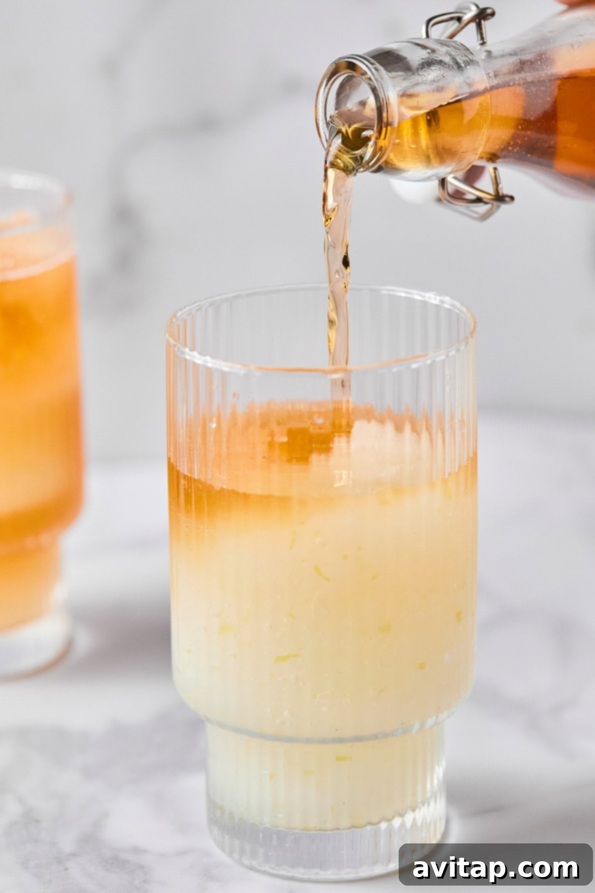 A glass containing a pale yellow, icy liquid is being filled with a darker orange liquid from a glass bottle, with another finished drink in the soft background.