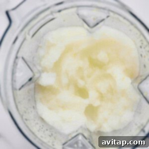 A top-down view of a blender containing a slushy, pale mixture with a slightly grainy texture on a white surface.