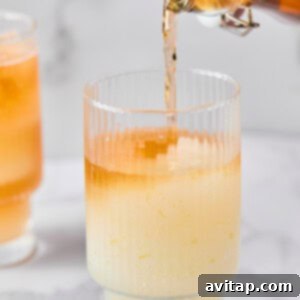 A glass with a pale yellow liquid and ice is being filled with an orange liquid from a glass bottle. Another similar drink is in the background.