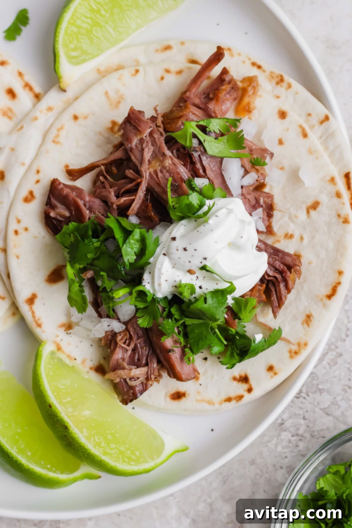 OLIPOP Infused Chuck Roast 2 A soft flour tortilla topped with generously shredded beef, finely chopped white onions, fresh green cilantro, and a dollop of creamy sour cream, served alongside vibrant lime wedges on a crisp white plate, showcasing a delicious taco.