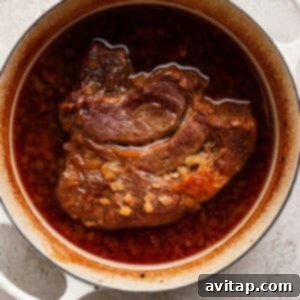 OLIPOP Infused Chuck Roast 12 A braised beef roast sits tenderly in a white Dutch oven, enveloped by a rich, red-brown broth and soft diced onions, all resting on a light textured surface, symbolizing hours of slow cooking.