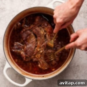 OLIPOP Infused Chuck Roast 13 A person skillfully uses two forks to shred succulent cooked beef within a round, white Dutch oven, surrounded by a rich, brown sauce, demonstrating the meat's perfect tenderness.