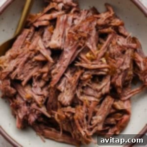 OLIPOP Infused Chuck Roast 14 A bowl filled with generously shredded cooked beef, glistening with its savory juices, with a fork gently resting on the side, inviting a taste.