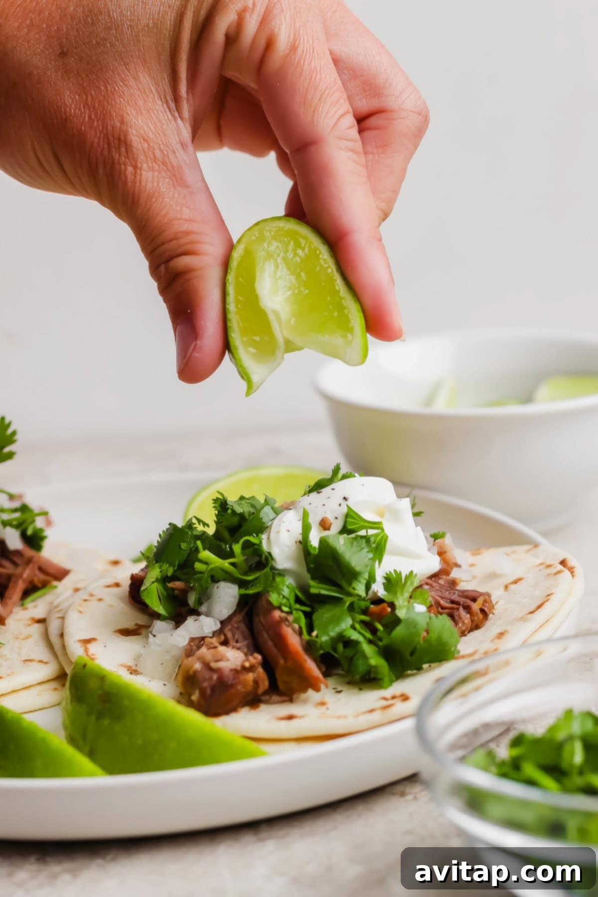 OLIPOP Infused Chuck Roast 6 A hand gently squeezes a fresh lime wedge over a perfectly assembled taco, generously filled with savory shredded meat, vibrant green cilantro, and a creamy dollop of sour cream, artfully placed on a pristine white plate. Additional lime wedges and a complementary bowl are subtly blurred in the background, creating an inviting culinary scene.
