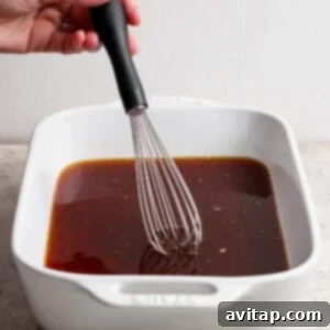 OLIPOP Infused Chuck Roast 8 A hand uses a whisk to mix a dark liquid marinade in a white rectangular baking dish on a light surface, preparing for the chuck roast.
