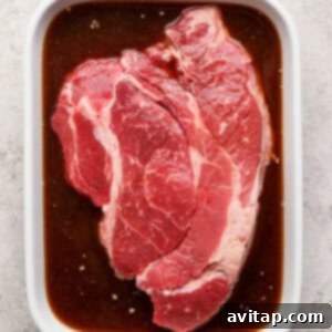 OLIPOP Infused Chuck Roast 9 A raw beef steak rests submerged in a rich, brown marinade within a white ceramic dish, positioned on a light textured surface, indicating the marination process.