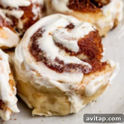 Close-up of a cinnamon roll with creamy white icing and swirls of cinnamon sugar on a light-colored plate.